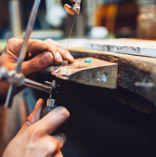 Primo piano di un gioielliere che usa un seghetto da traforo per tagliare un piccolo pezzo di metallo su un banco da lavoro in legno.