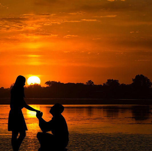 Uomo in ginocchio sulla spiaggia al tramonto, propone matrimonio con anello di diamanti, donna sorpresa felice. Scena romantica proposta di matrimonio a Roma con anello di fidanzamento.